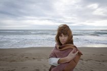 Donna sulla spiaggia avvolta in una coperta guardando la macchina fotografica — Foto stock