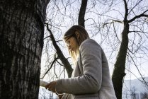 Young woman standing beside tree, reading book, low angle view — Stock Photo