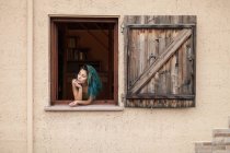 Молода жінка з блакитним волоссям, дивиться з відкритого вікна — стокове фото