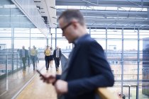 Geschäftsmann schaut aufs Smartphone auf Bürobalkon, Menschen gehen im Hintergrund — Stockfoto