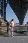 Mujer joven haciendo ejercicio al aire libre, estirándose, debajo del puente Williamsburg, Nueva York, EE.UU. - foto de stock