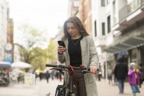 Woman browsing mobile phone while standing with bicycle in city street — Stock Photo