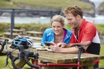 Cyclists using mobile phone at picnic table overlooking ocean — Stock Photo