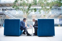 Два бизнесмена обсуждают встречу в офисных креслах атриума — стоковое фото