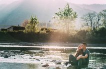 Pareja romántica sentada sobre rocas junto al río, sonriendo - foto de stock