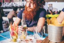 Joven hipster masculino con pelo rojo y barba bebiendo cerveza embotellada en el bar de la acera - foto de stock
