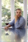 Homem em casa, sentado à mesa, segurando um copo de vinho, expressão pensativa — Fotografia de Stock