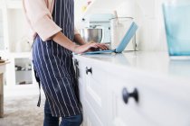 Обрезанное изображение пекаря-женщины на кухонном столе, печатающего на ноутбуке — стоковое фото
