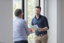 Male colleagues at office window standing and talking while holding paper documents — Stock Photo