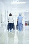Vista trasera de los camilleros femeninos empujando sillas de ruedas a lo largo del pasillo del hospital - foto de stock