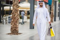 Uomo che indossa abiti tradizionali mediorientali passeggiando lungo la strada portando borse della spesa, Dubai, Emirati Arabi Uniti — Foto stock