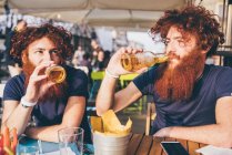 Молоді чоловіки хіпстер близнюки з рудим волоссям і бородою п'ють пиво на тротуарі — стокове фото