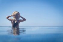 Rear view of young woman standing in blue sea with hands on sunhat, Villasimius, Sardinia, Italy — Stock Photo