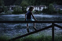 Niño sentado en la valla por el río mirando por encima del hombro a la cámara - foto de stock