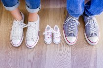 Cropped view of baby shoes between male and female feet — Stock Photo