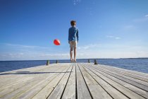 Young boy jumping on wooden pier, holding red helium balloon, rear view — Stock Photo