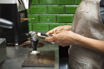 Sección media de mensajes de texto barista femenina en el teléfono inteligente en la cocina cafetería - foto de stock