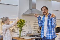 Daughter taking photograph of funny-faced father in kitchen — Stock Photo