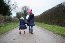 Vista trasera de niña y niña en sombreros de punto caminando por el camino rural - foto de stock