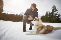 Junger Mann hockt mit Husky in verschneiter Landschaft, Elmau, Bayern, Deutschland — Stockfoto