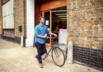 Young man carrying coffee and wheeling pushbike past workshop — Stock Photo