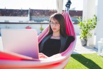 Young woman reclining in city roof terrace hammock typing on laptop — Stock Photo