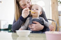 Mulher adulta média alimentando filha bebê na mesa da cozinha — Fotografia de Stock