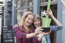 Mujeres frente a la tienda sosteniendo letrero abierto tomando selfie con teléfono inteligente - foto de stock