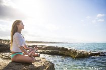 Young woman sitting on rocks by sea, in yoga position — Stock Photo