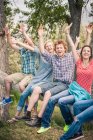 Portrait of teenage girl and young adult friends sitting on fallen tree with hands raised — Stock Photo