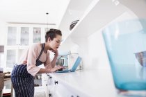 Молода жінка пекарня спирається на кухонну лічильницю, дивлячись на ноутбук — стокове фото