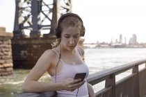 Mujer joven al lado del río, con auriculares, usando smartphone, Nueva York, EE.UU. - foto de stock