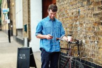 Junger Mann hält Kaffee und checkt Handy, schiebt Fahrrad gegen Ziegelwand — Stockfoto