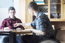Молодой человек в инвалидном кресле играет в шашки с другом на кухне — стоковое фото