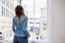 Vue arrière de la jeune femme écoutant de la musique regardant par la fenêtre de l'appartement de la ville — Photo de stock