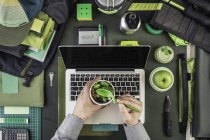 Cropped image of man holding salad whilst preparing hiking equipment and laptop — Stock Photo