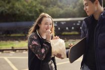 Hombre joven compartiendo bolsa de papas fritas con mujer joven, monopatín bajo el brazo de hombre joven, Bristol, Reino Unido - foto de stock
