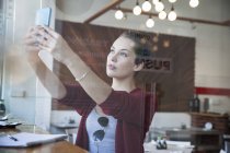 Молода жінка сидить у кафе, приймає селфі, використовуючи смартфон — стокове фото