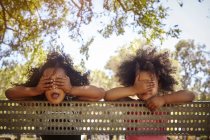 Portrait of two young sisters leaning against fence, covering eyes — Stock Photo