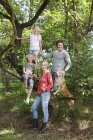 Семья в лесу лазает по дереву глядя на камеру улыбаясь — стоковое фото