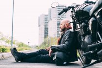 Älterer männlicher Motorradfahrer sitzt am Straßenrand und lehnt sich mit Smartphone an Motorrad — Stockfoto