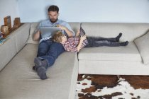 Niño acostado en el sofá con el padre utilizando la tecnología - foto de stock