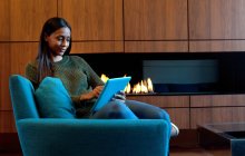 Woman in armchair in front of wooden cabinets and open fire, using digital tablet — Stock Photo
