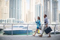 Pareja paseando por el paseo marítimo llevando bolsas de compras, Dubai, Emiratos Árabes Unidos - foto de stock