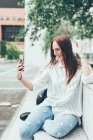 Молода жінка сидить на стіні, приймаючи смартфон селфі — стокове фото