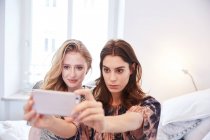Two young women sitting on bed taking smartphone selfie — Stock Photo