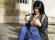Junge Frau sitzt neben Säule, hält Kaffeetasse zum Mitnehmen in der Hand, schaut aufs Smartphone — Stockfoto