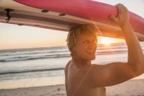 Чоловік на пляжі носить дошку для серфінгу над головою, дивлячись через плече на камеру посміхаючись — стокове фото