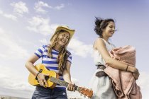 Duas jovens mulheres jogando ukulele e carregando cobertor contra o céu azul — Fotografia de Stock
