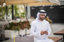 Hombre comprador con ropa tradicional de Oriente Medio sentado en el banco de lectura de texto smartphone, Dubai, Emiratos Árabes Unidos - foto de stock